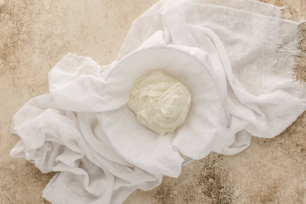 A bowl of thick, creamy yogurt sits on a white cheesecloth, which is draped across a textured beige surface.