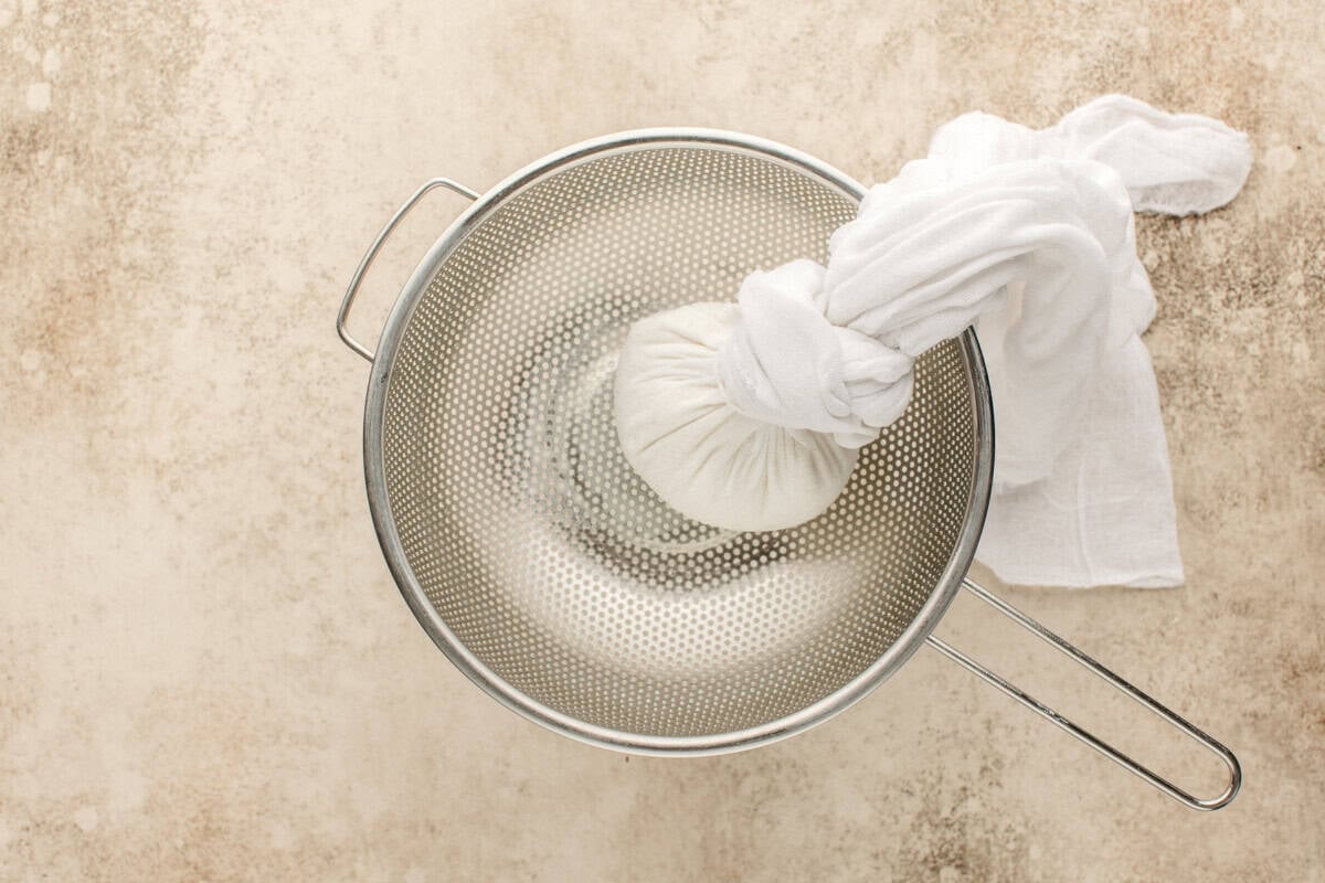 A metal strainer with a handle is placed on a beige surface. Inside the strainer, a bundle of white cloth is tied at the top around salted greek yogurt being strained.