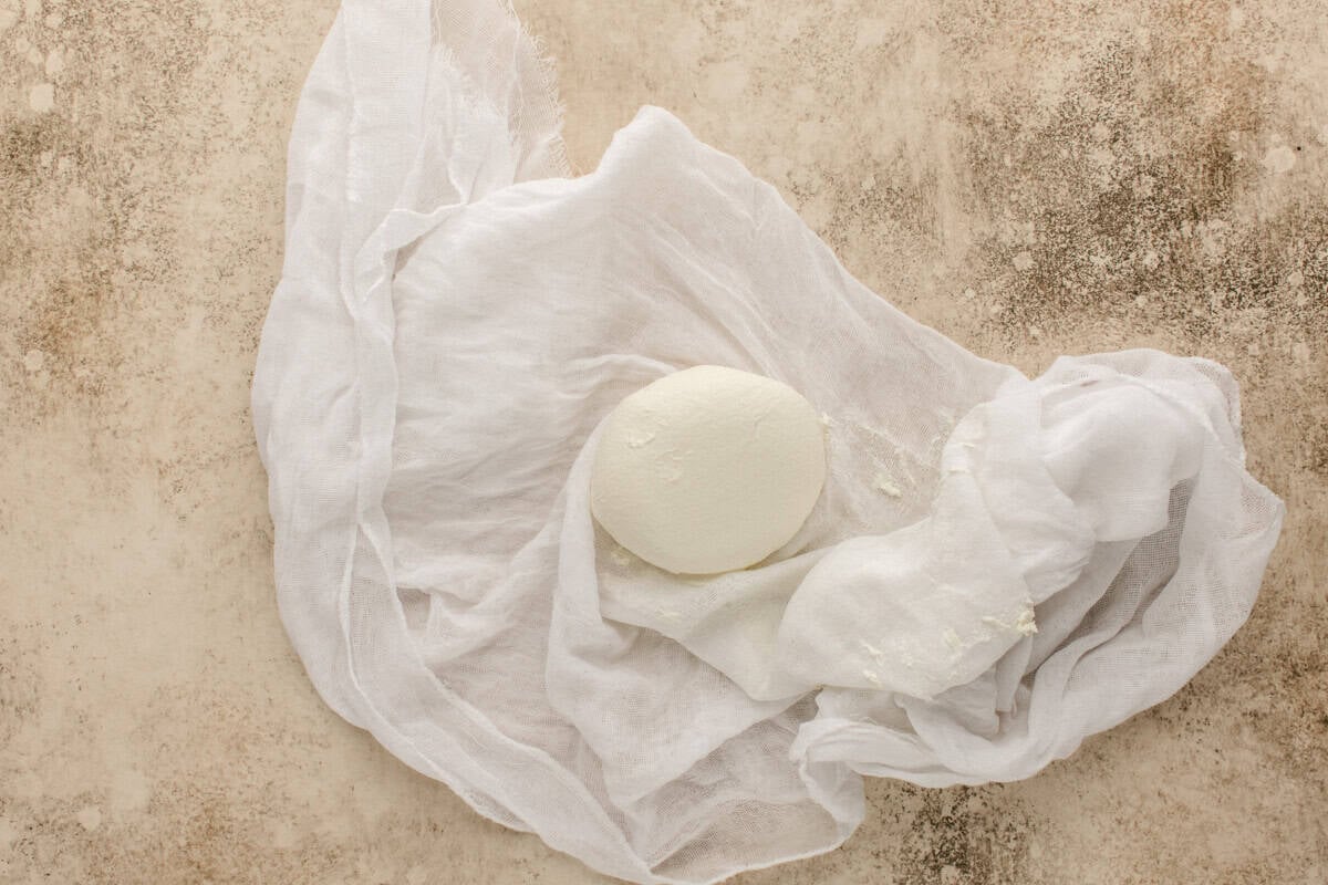 A round piece of fresh, white labneh sits on a loosely draped piece of cheesecloth, placed on a speckled beige surface.