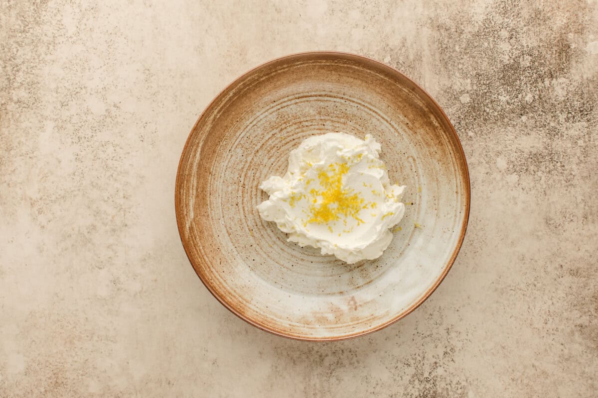 A brown-rimmed ceramic plate holds labneh topped with grated lemon zest, set on a textured beige surface.