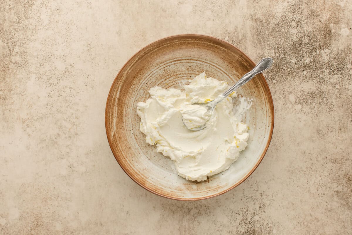 A brown ceramic bowl with creamy labneh and lemon zest being stirred by a silver spoon, placed on a textured beige surface.