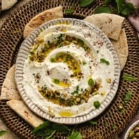 A bowl of creamy whipped labneh dip with chopped pistachios, fresh herbs, olive oil, and spices, surrounded by pita bread slices on a woven tray.