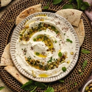 A bowl of creamy whipped labneh dip with chopped pistachios, fresh herbs, olive oil, and spices, surrounded by pita bread slices on a woven tray.
