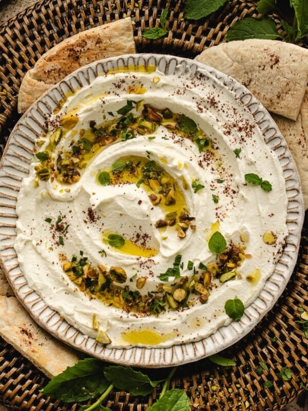A bowl of creamy whipped labneh dip with chopped pistachios, fresh herbs, olive oil, and spices, surrounded by pita bread slices on a woven tray.