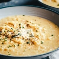 A close-up of a bowl of creamy pastina soup, topped with shredded cheese, pepper, and herbs, set on a table. The text above and below reads "Pinch and Swirl Pastina Recipe" and "pinchandswirl.com".