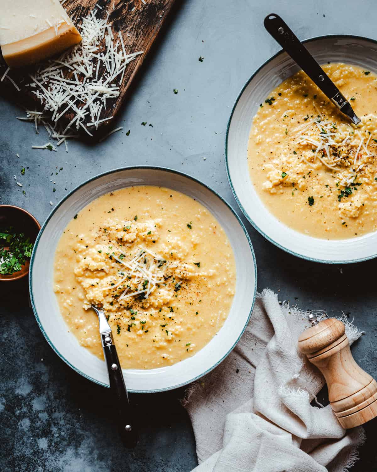 Two bowls of creamy pastina topped with shredded cheese and chopped herbs, placed on a gray surface with spoons. Nearby are a wooden board with grated cheese, a small bowl of herbs, a napkin, and a wooden pepper grinder.