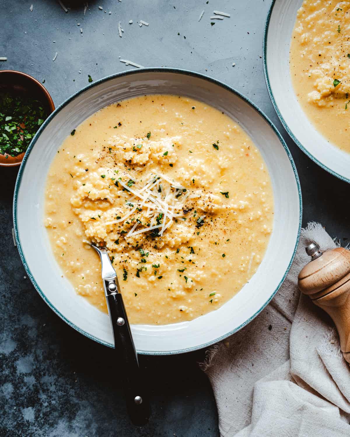 A bowl of creamy pastina topped with grated cheese, fresh herbs, and black pepper, with a spoon resting inside. Nearby are a pepper mill, a beige napkin, and a small bowl of chopped herbs.