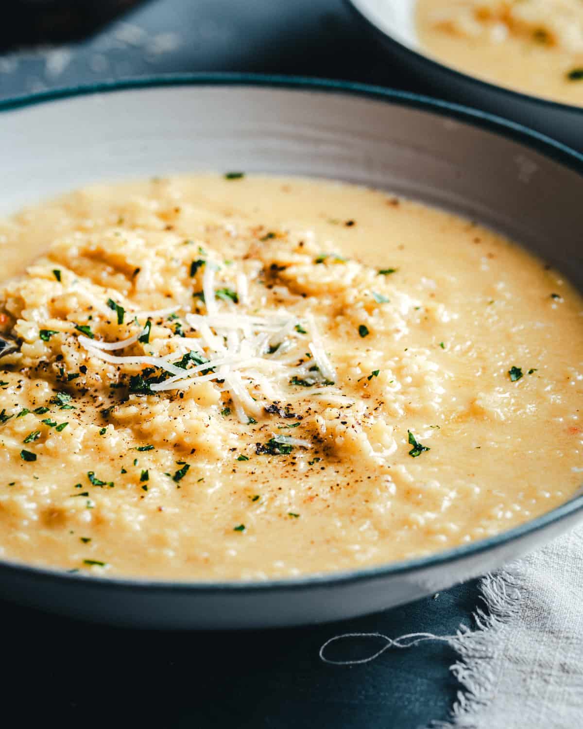 A close-up of a creamy bowl of pastina soup, garnished with shredded cheese, chopped herbs, and cracked black pepper, served in a gray and white bowl on a dark surface.
