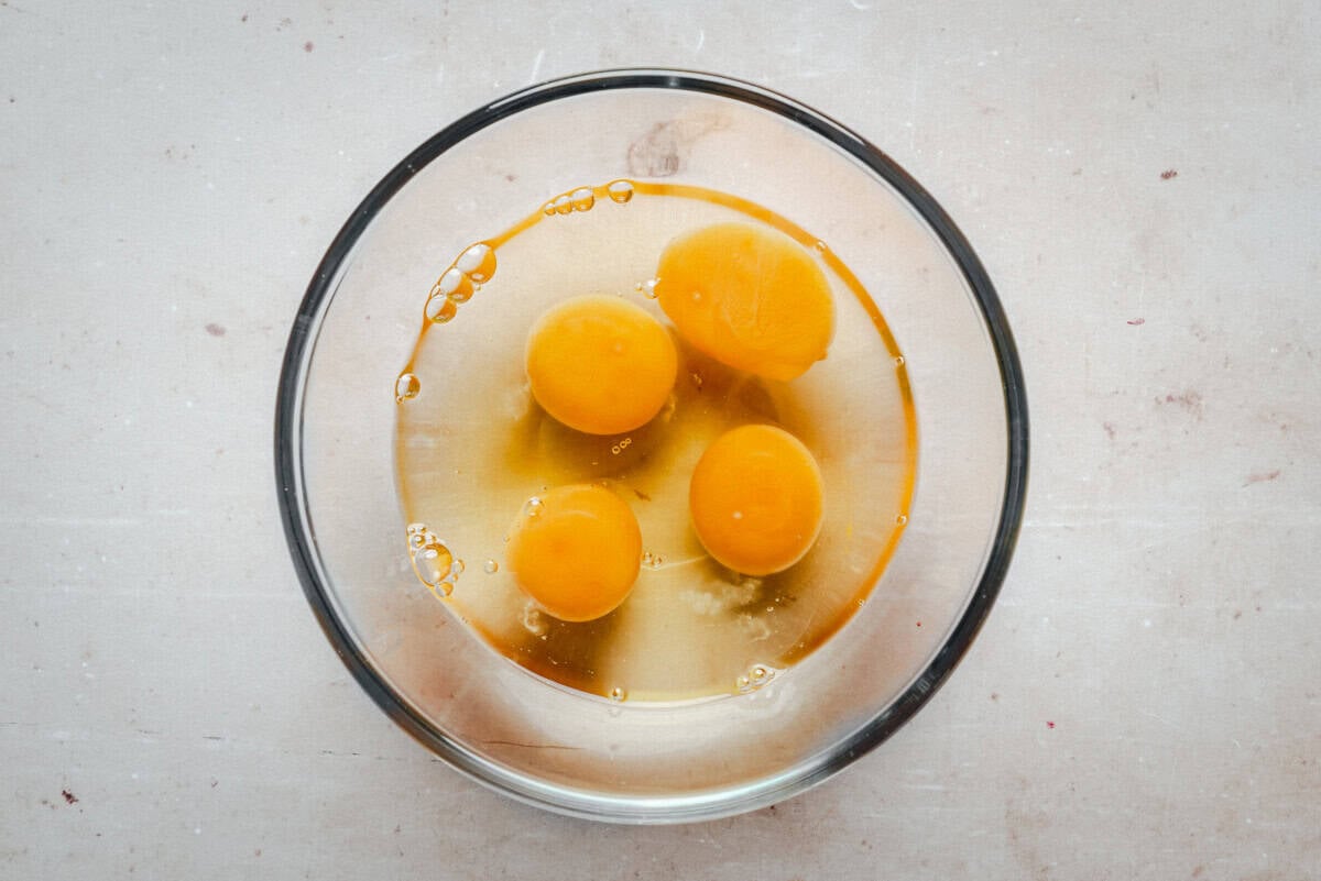 A glass bowl containing four raw eggs with their yolks and whites visible, placed on a light-colored countertop.