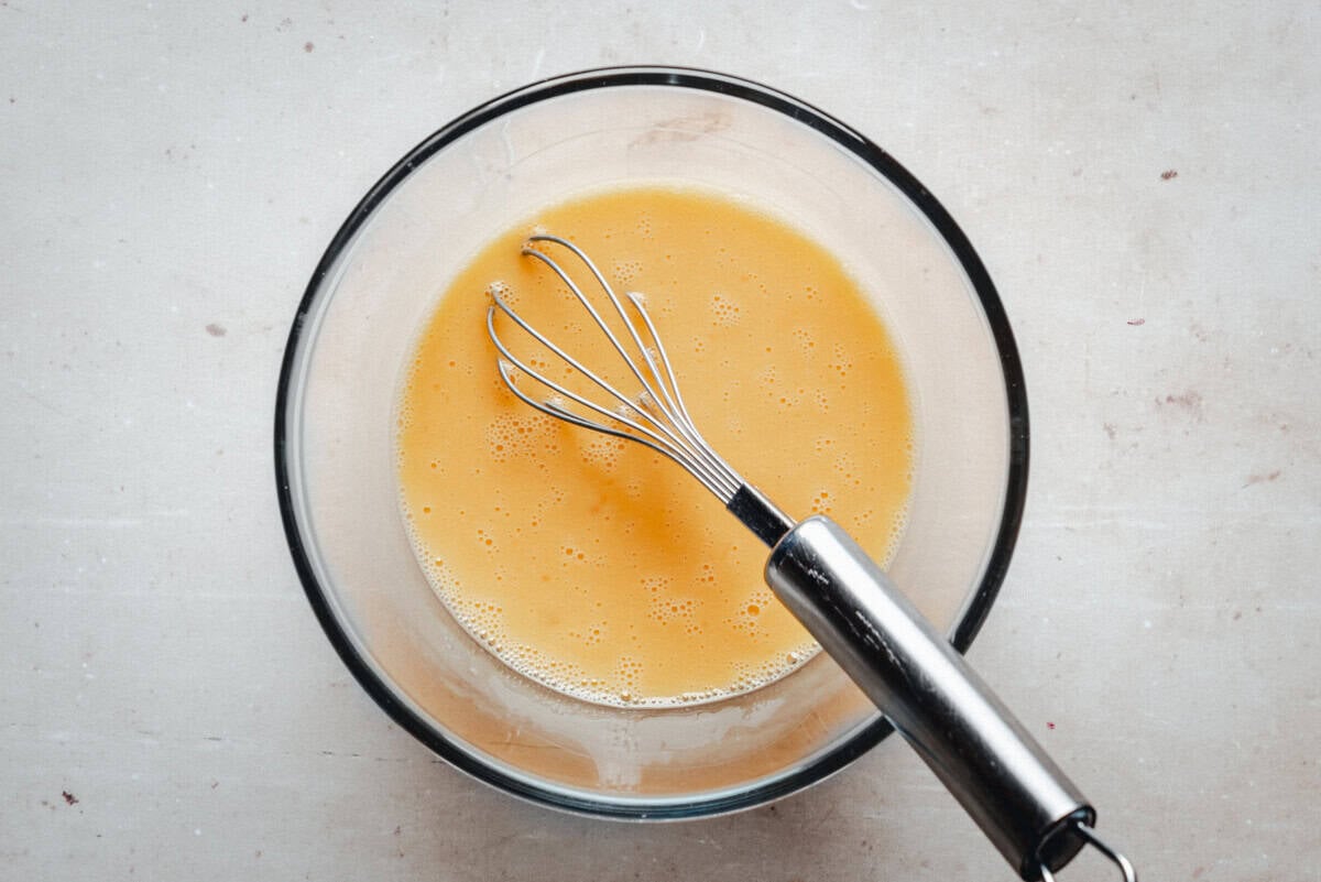 A metal whisk rests in a glass bowl filled with beaten eggs, set on a light, speckled surface.