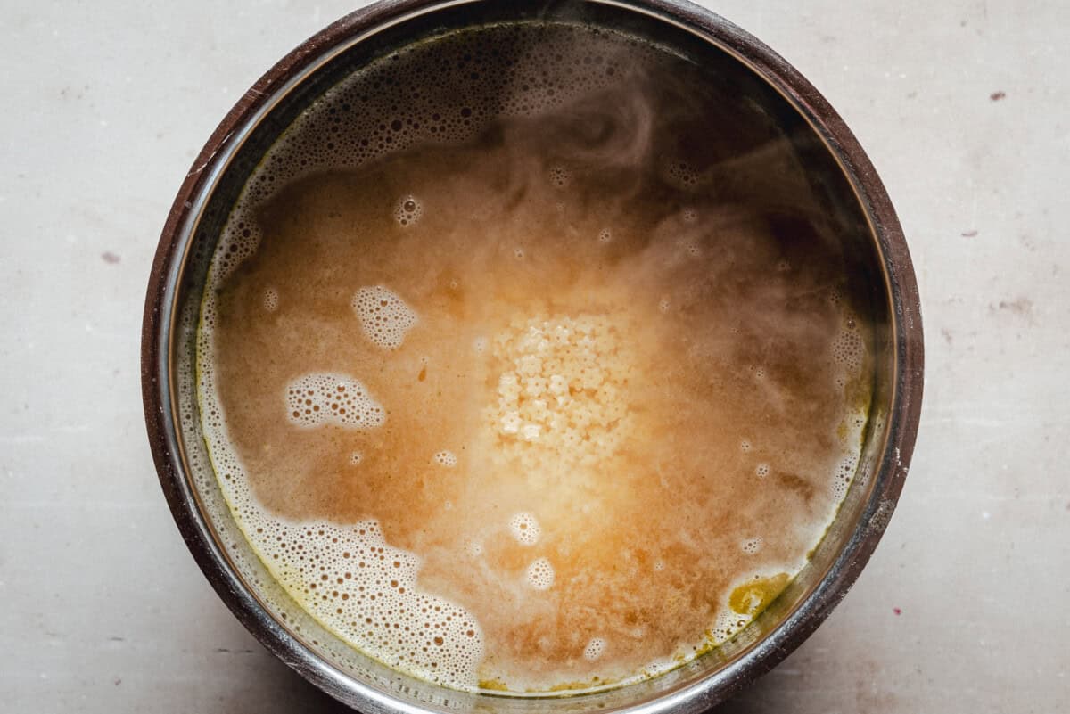 A metal pot filled with hot, steaming broth and small pastina pasta cooking in the center, surrounded by bubbles and foam on the surface.