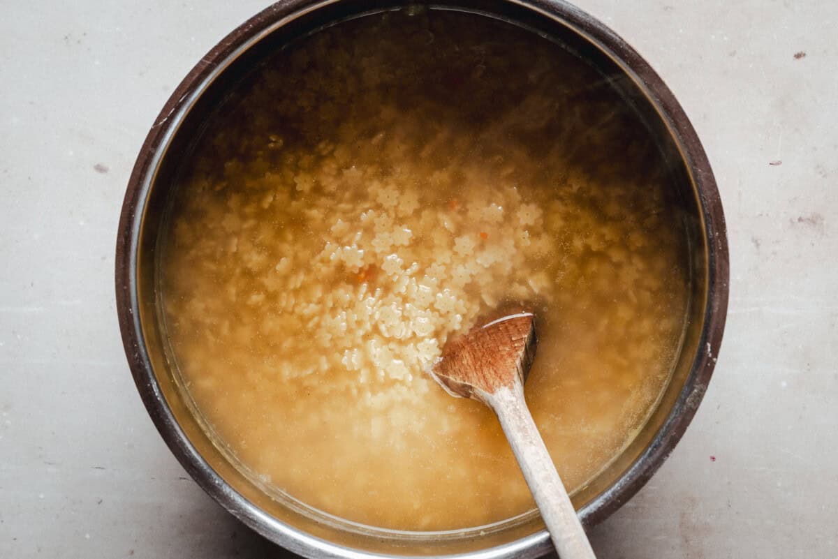 A pot of soup with small star-shaped pasta and a wooden spoon resting inside, viewed from above. The soup appears to be broth-based and is light in color.