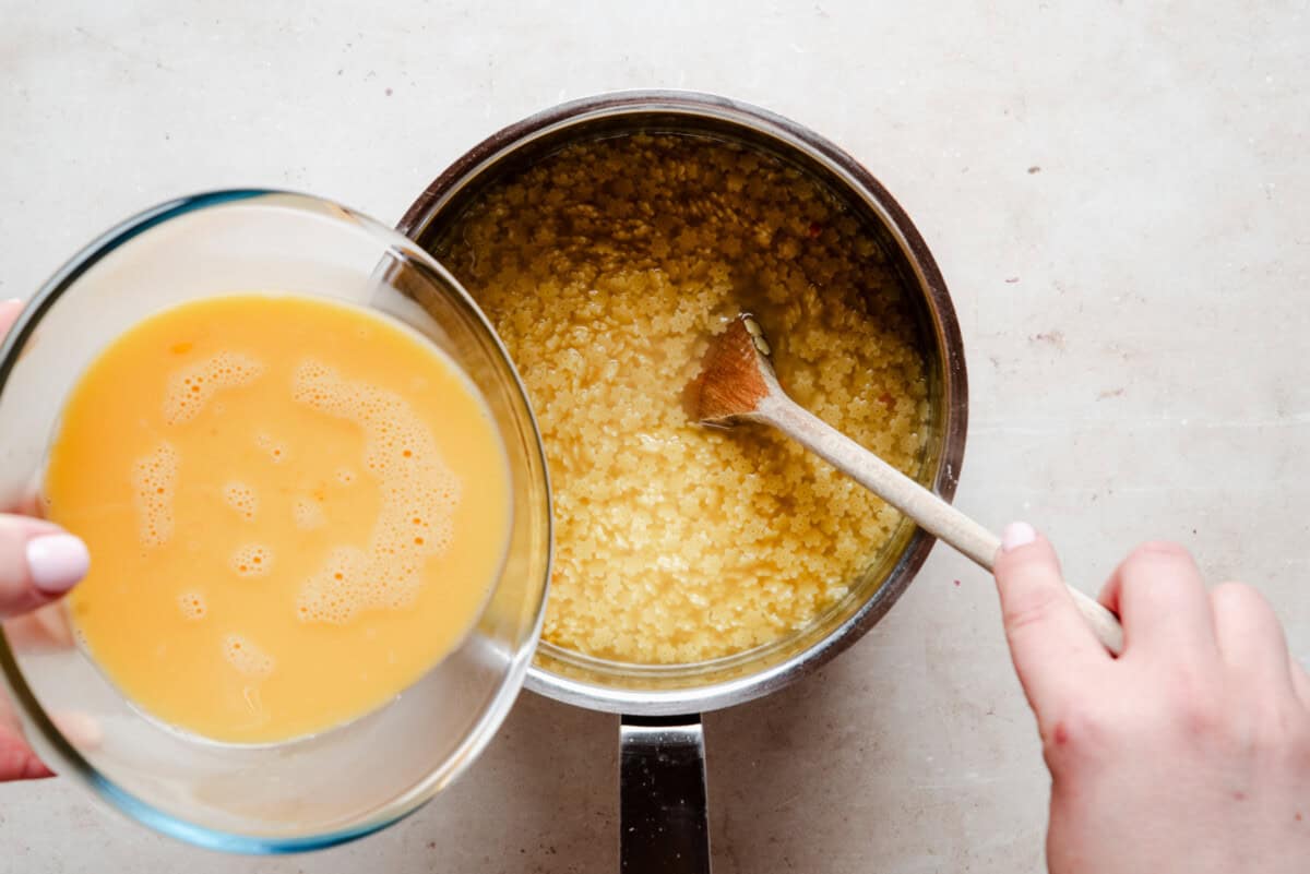 A person pours a bowl of beaten eggs into a saucepan of cooked pastina pasta, stirring with a wooden spoon on a light countertop.