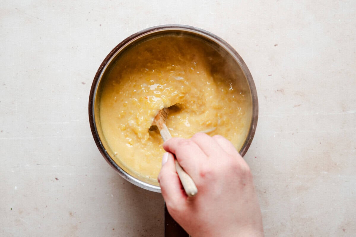 A hand stirs beaten eggs into pastina with a wooden spoon in a metal saucepan placed on a light-colored countertop.