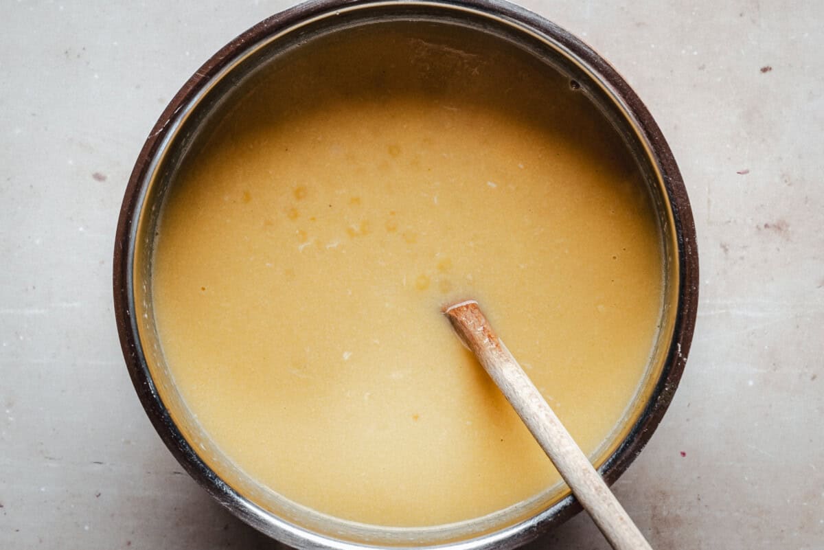 A round brown pot filled with creamy broth, with a wooden spoon resting inside, viewed from above on a light surface.