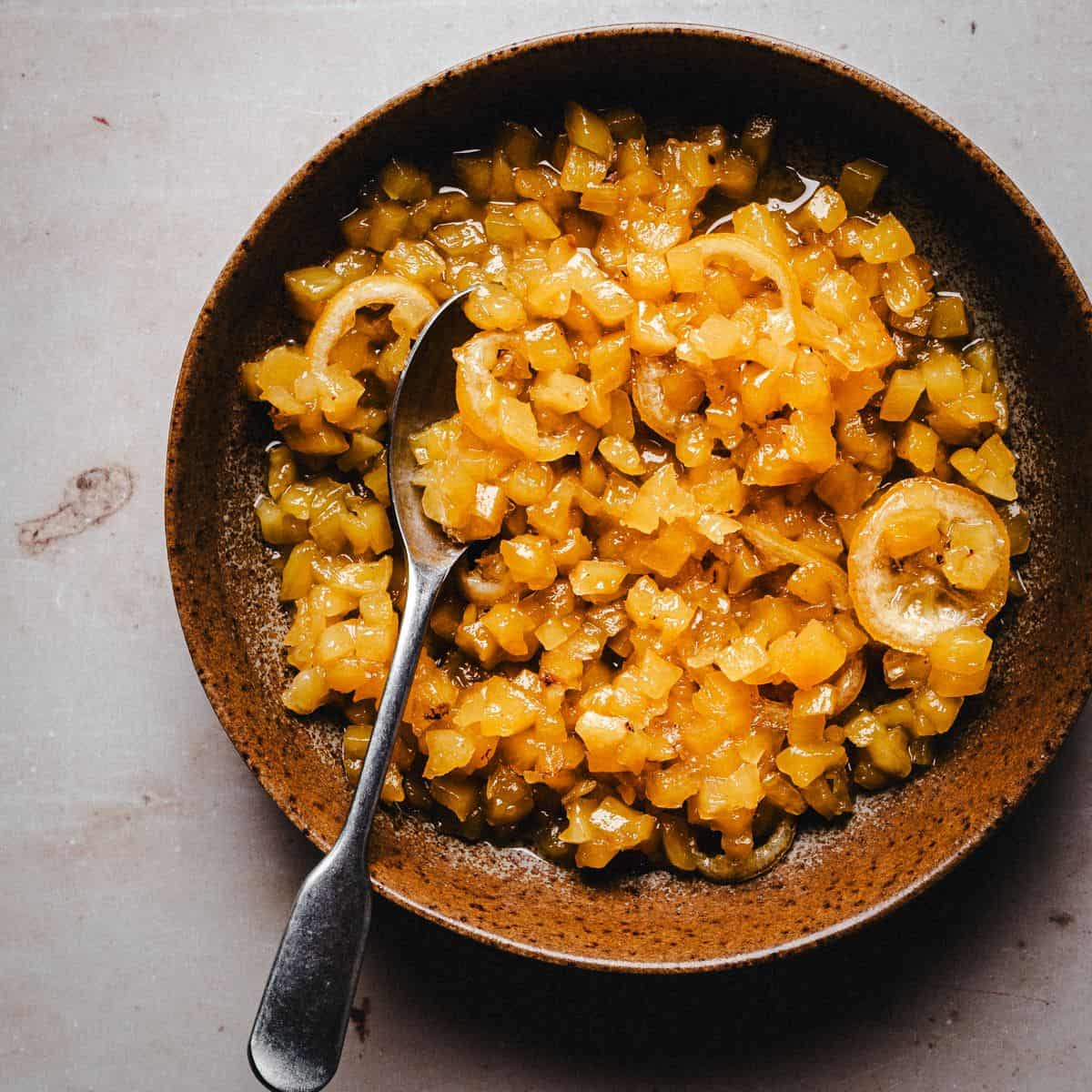 A brown bowl filled with pineapple marmalade with a silver spoon resting inside, placed on a light-colored surface.