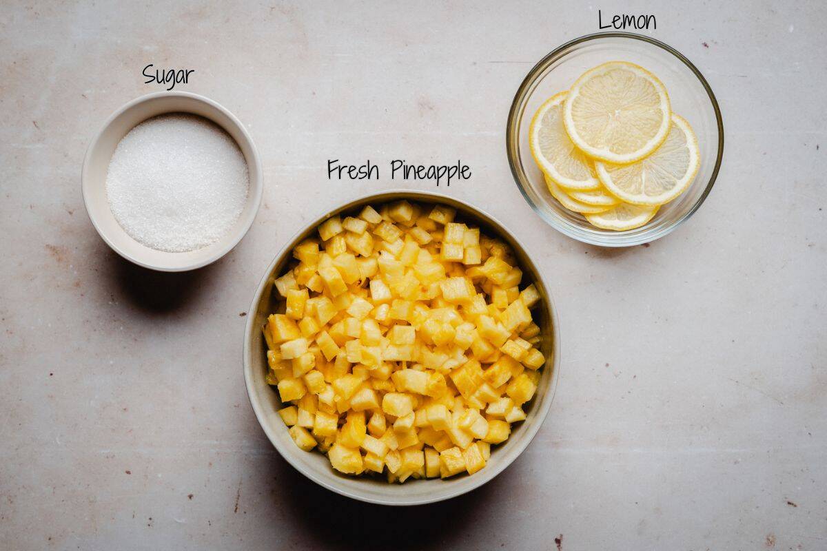 A bowl of diced fresh pineapple, a bowl of sugar, and a bowl of sliced lemon on a light surface, each labeled accordingly.