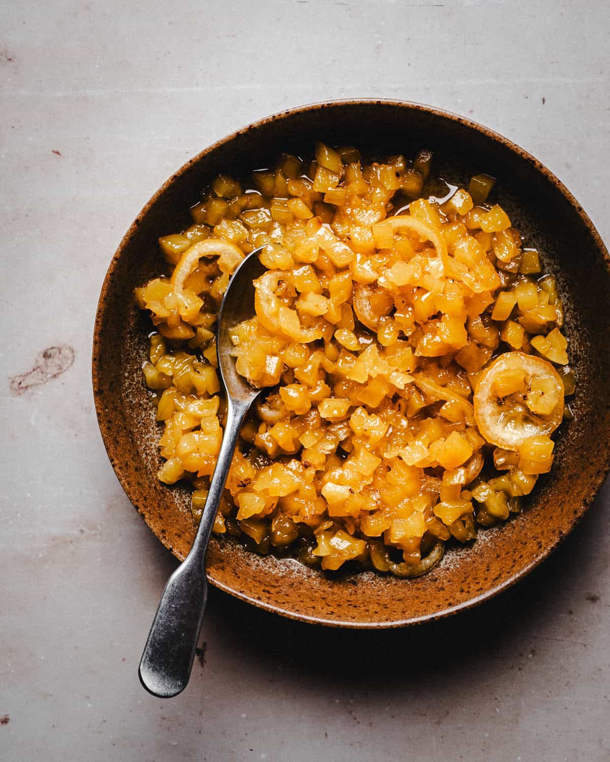 A brown ceramic bowl filled with pineapple marmalade. A metal spoon rests inside the bowl on a light, textured surface.