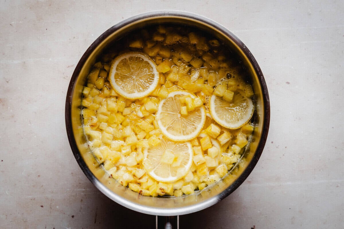 A saucepan filled with diced pineapple and several round lemon slices in liquid, viewed from above on a light-colored surface.
