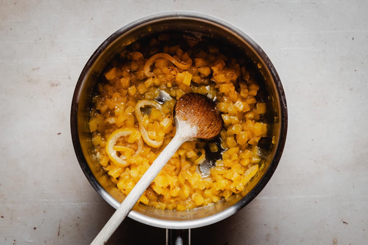A metal pot filled with pineapple marmalade, being stirred with a wooden spoon on a light-colored surface.
