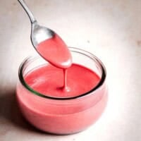 A spoon drizzles pink raspberry vinaigrette into a small glass jar on a light surface. Text above reads "Raspberry Vinaigrette" and "pinchandswirl.com" is written below.
