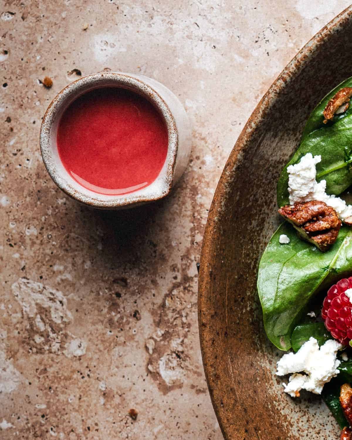 A small pitcher of raspberry vinaigrette sits next to a rustic brown plate containing spinach leaves, feta cheese, pecans, and a raspberry, on a textured stone surface.