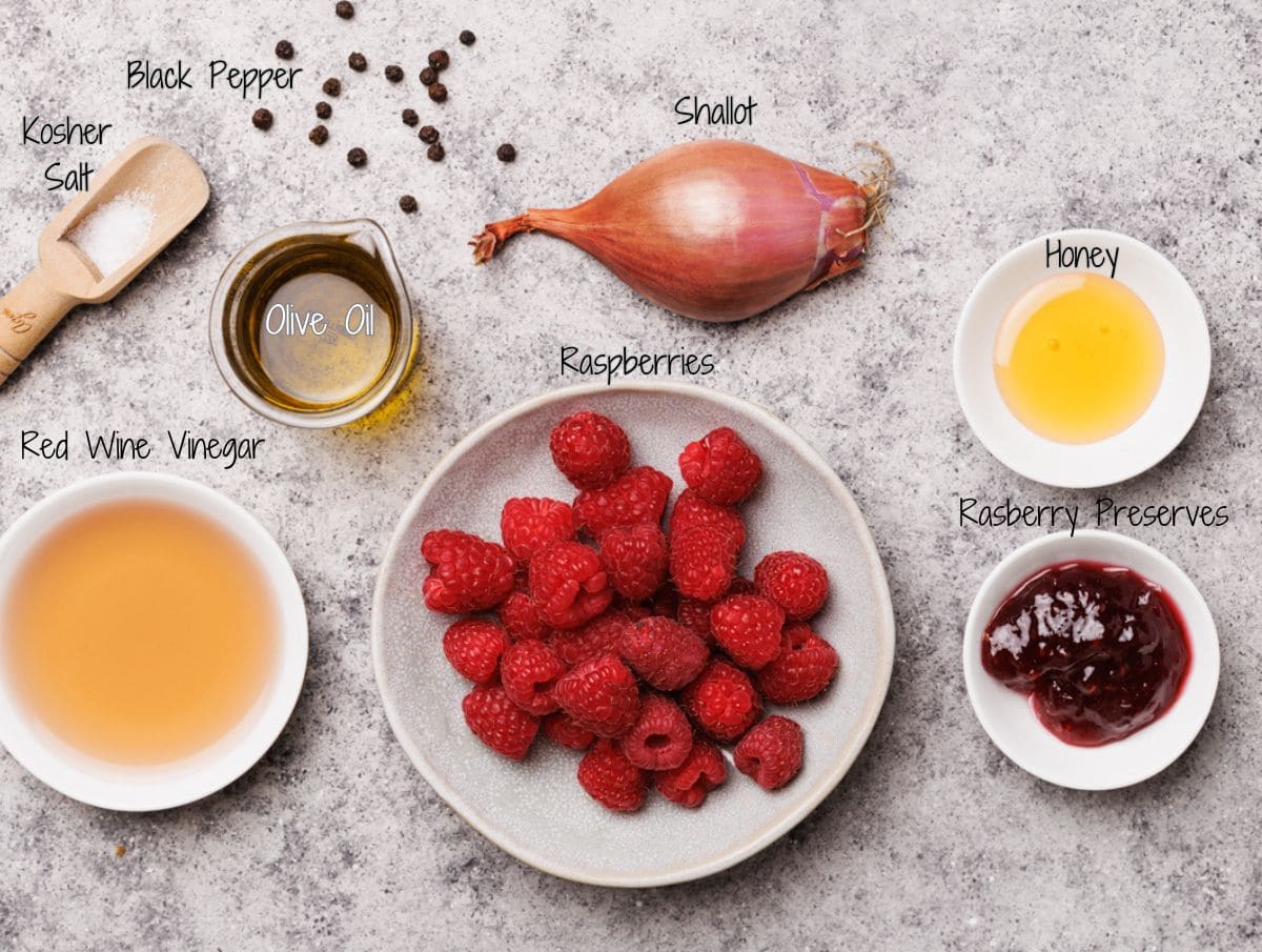 A bowl of raspberries surrounded by small bowls of olive oil, red wine vinegar, honey, raspberry preserves, a shallot, and a wooden spoon of kosher salt with black peppercorns on a stone surface.