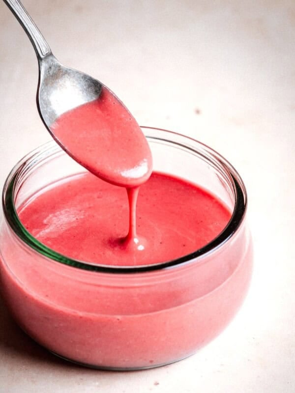 A spoon drizzles pink, creamy raspberry vinaigrette into a small glass jar filled with the same sauce, set on a light beige surface.