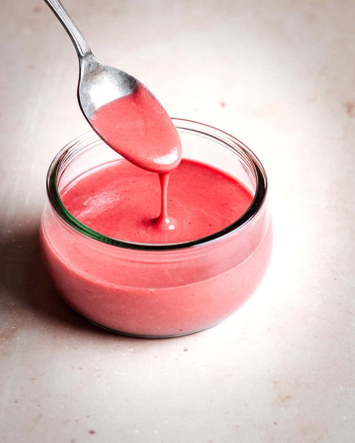 A spoon drizzles raspberry vinaigrette into a small glass jar on a light, textured surface.