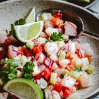 A bowl of shrimp ceviche garnished with lime wedges, cilantro, and diced vegetables like red onion, cucumber, and tomato, with a spoon resting inside the bowl.