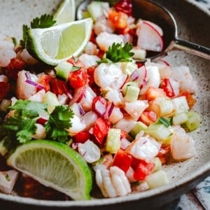 A close-up of shrimp ceviche with diced red onions, tomatoes, cucumbers, and cilantro, garnished with lime wedges, served in a speckled bowl with a spoon.