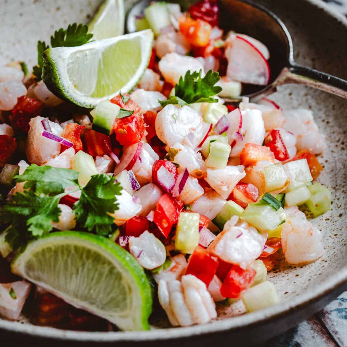 A close-up of shrimp ceviche with diced red onions, tomatoes, cucumbers, and cilantro, garnished with lime wedges, served in a speckled bowl with a spoon.