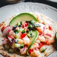 A shrimp ceviche tostada topped with diced shrimp, cucumber, tomato, red onion, microgreens, and avocado slices on a tortilla, served on a decorative plate with a lime wedge.