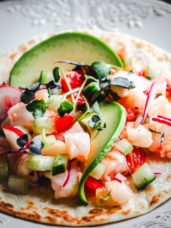 A close-up of a shrimp ceviche tostada on a white plate, topped with avocado slices, radish, cucumber, microgreens, and tomato. A lime wedge is visible on the side.