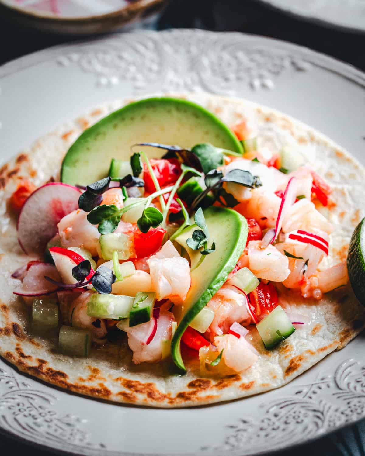 Shrimp ceviche tostada topped with diced shrimp, avocado slices, microgreens, radish, cucumber, and tomato.