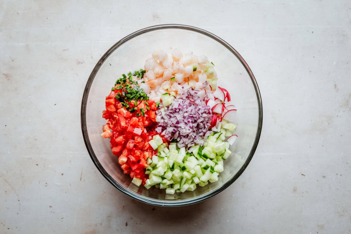 Shrimp ceviche with diced cucumber, red onion, tomato, radish, and fresh herbs in a glass bowl.