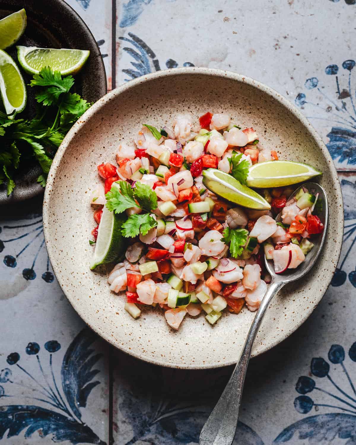 A bowl of shrimp ceviche with diced shrimp, tomato, cucumber, and red onion, garnished with fresh cilantro and lime wedges. A spoon rests in the bowl, with lime wedges and cilantro on a plate nearby. The bowl sits on a patterned surface.