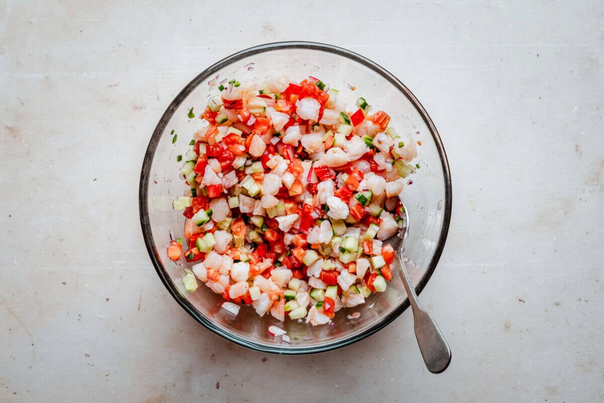 A glass bowl filled with diced shrimp, tomatoes, cucumbers, onions, and herbs, with a spoon resting inside, sits on a light-colored surface.