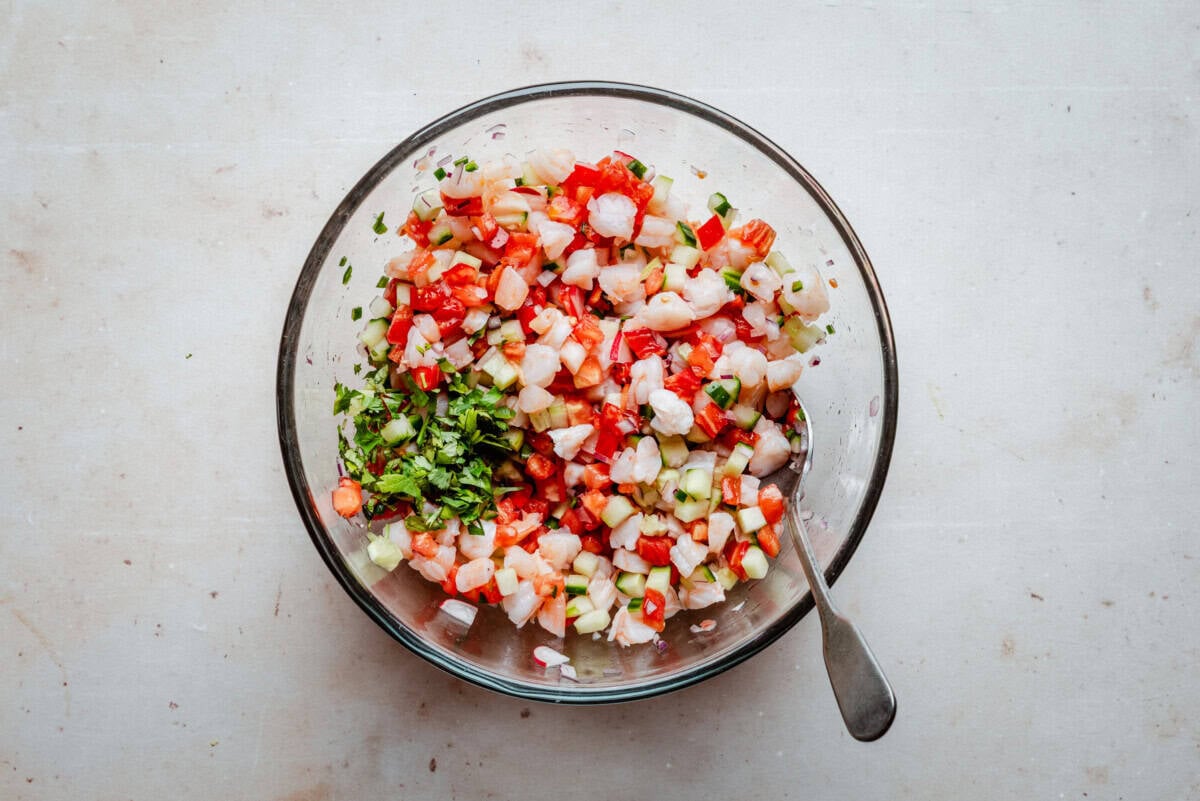 A glass bowl filled with ceviche made of diced shrimp, tomatoes, onions, cucumbers, and cilantro, with a spoon resting inside. The bowl sits on a light-colored surface.