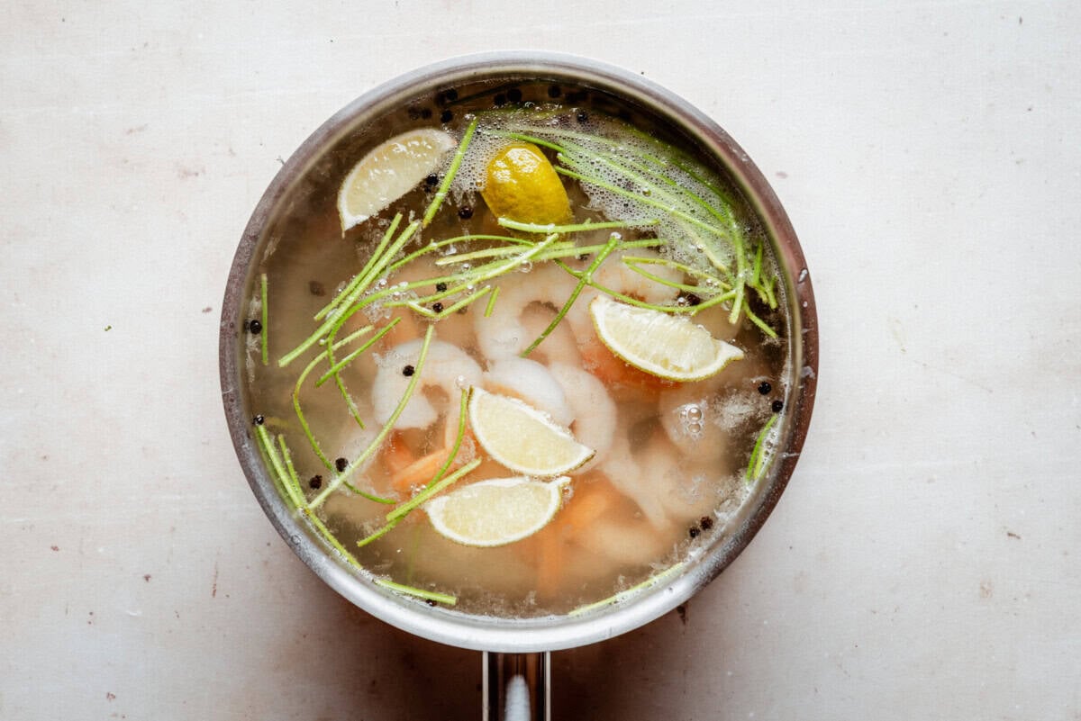A pot of broth with shrimp, lemon wedges, green herbs, and black peppercorns floating on the surface, viewed from above on a light background.