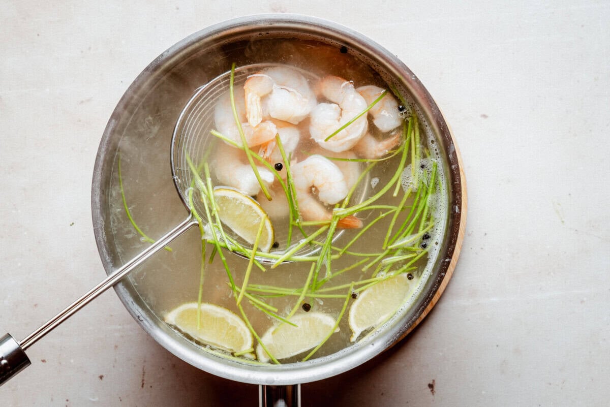 A pot of clear soup with shrimp, lime wedges, green herbs, and black peppercorns. A slotted spoon is lifting some shrimp from the broth. The pot is on a light-colored surface.