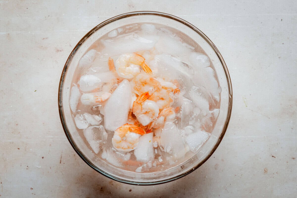 A glass bowl filled with shrimp and ice sits on a light-colored surface, with the shrimp partially submerged in water and surrounded by ice cubes.