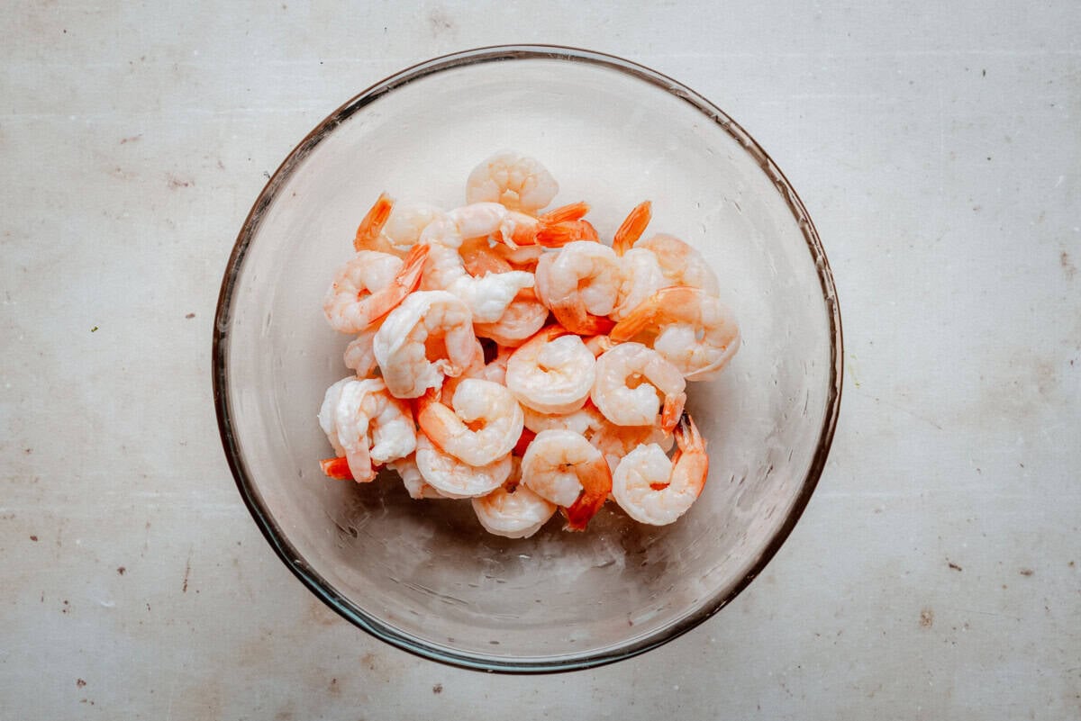 A glass bowl filled with cooked, peeled shrimp is placed on a light-colored countertop.