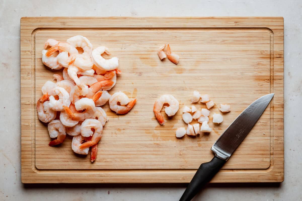 A wooden cutting board with shrimp—some whole, some chopped—next to a small kitchen knife with a black handle. The board is placed on a light-colored surface.