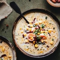 A bowl of creamy shrimp and corn bisque, garnished with shrimp, corn kernels, bacon bits, and herbs, with a spoon resting in the bowl. Another partial bowl and a napkin are visible on a rustic green surface.