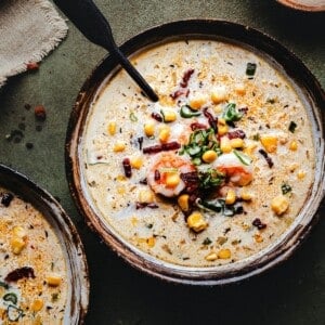 A bowl of creamy shrimp and corn bisque topped with shrimp, corn, chopped green onions, and bacon bits, garnished with herbs and spices. A black spoon rests in the bowl, and part of another bowl is visible nearby.