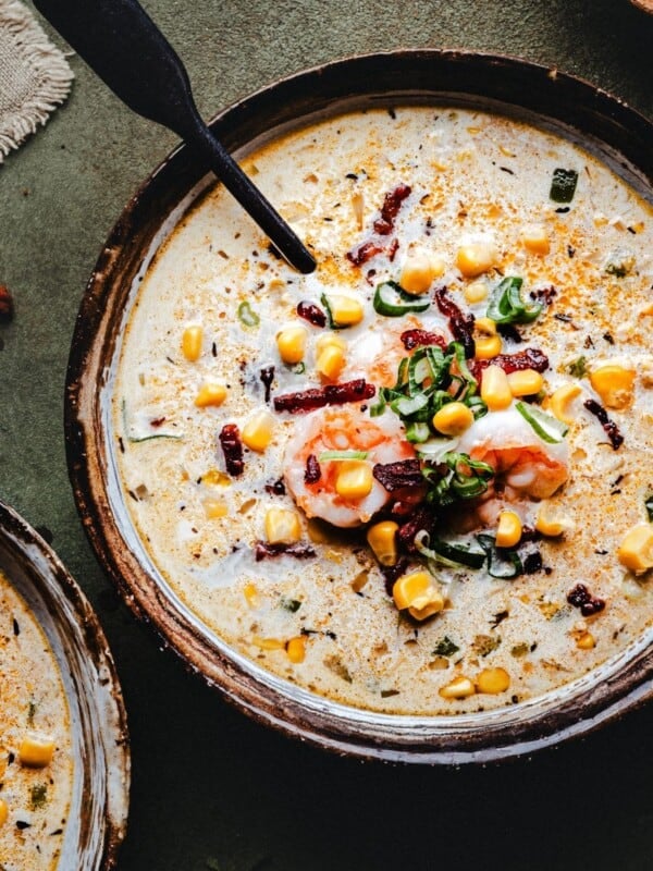 A bowl of creamy shrimp and corn bisque topped with shrimp, corn, chopped green onions, and bacon bits, garnished with herbs and spices. A black spoon rests in the bowl, and part of another bowl is visible nearby.