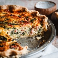 A baked spinach bacon quiche in a pie pan with a slice missing, showing the creamy filling with spinach and bacon. The edge of the crust is golden brown. Text at the top reads "Spinach Bacon Quiche.