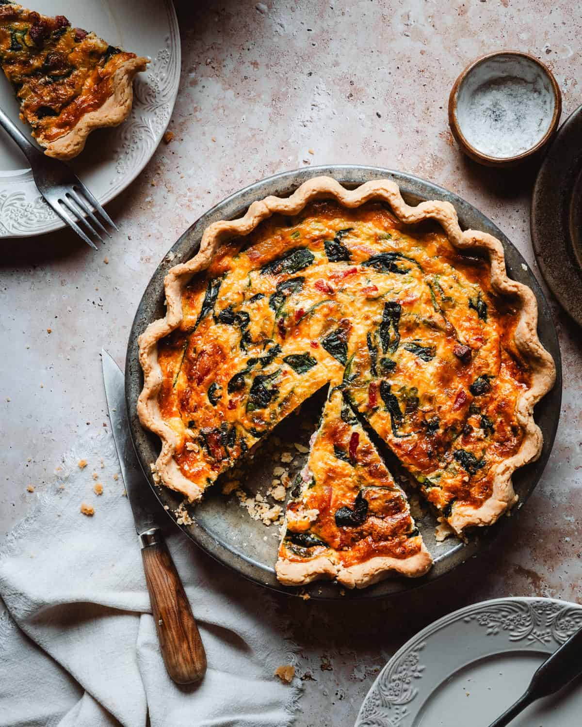 A baked spinach bacon quiche with a golden crust, filled with spinach and cheese, sits on a textured surface. One slice is being removed, and plates, a knife, and a white napkin are nearby.