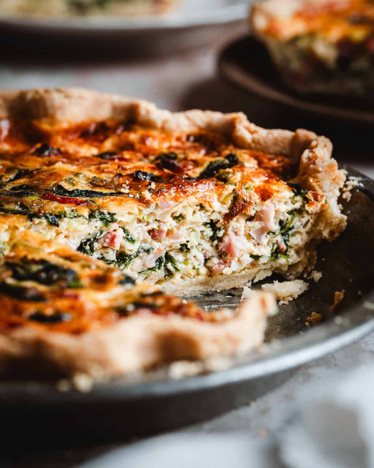 A close-up of a baked spinach bacon quiche with a golden crust, filled with spinach, ham, and cheese. A slice has been removed, revealing the creamy filling and flaky pastry inside a metal pie dish.